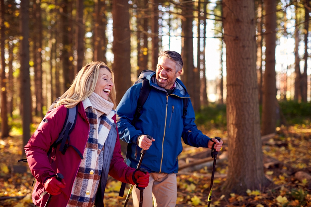 A smiling man and woman with backpacks walk through a sunlit forest with hiking poles. They wear jackets and warm clothes, enjoying a pleasant hike among tall trees and fallen autumn leaves.