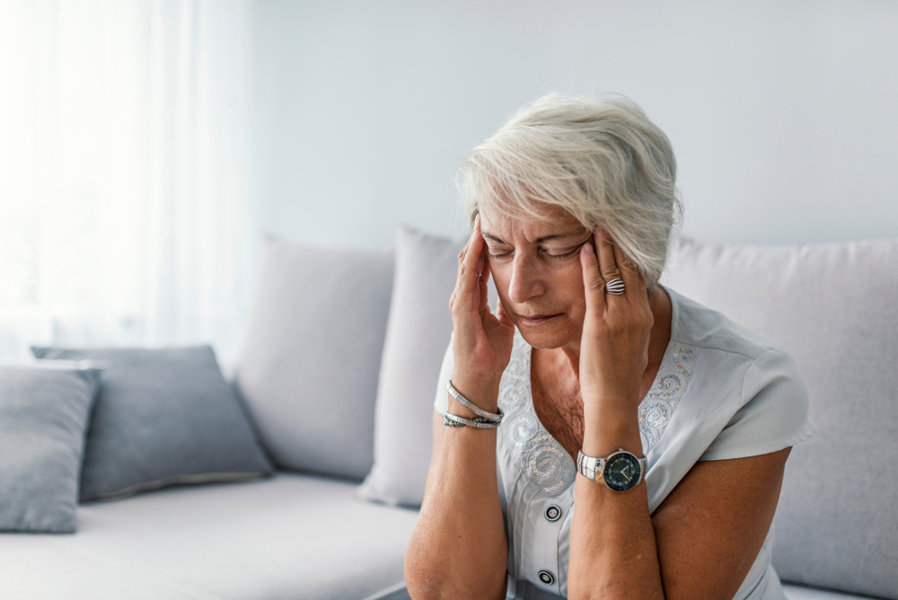 An elderly woman with short gray hair sits on a couch, holding her temples and closing her eyes, appearing to be in pain or discomfort. She wears a watch and jewelry, with a concerned expression on her face.