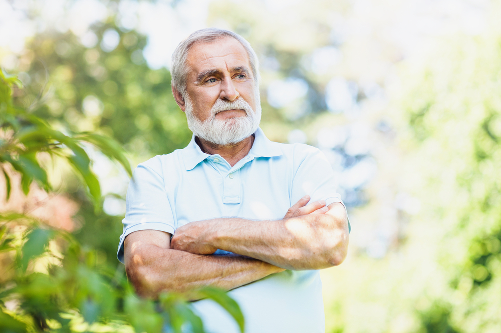 An older man with white hair and a beard stands outdoors with arms crossed, wearing a light blue polo shirt. He looks thoughtful, surrounded by greenery and soft sunlight.