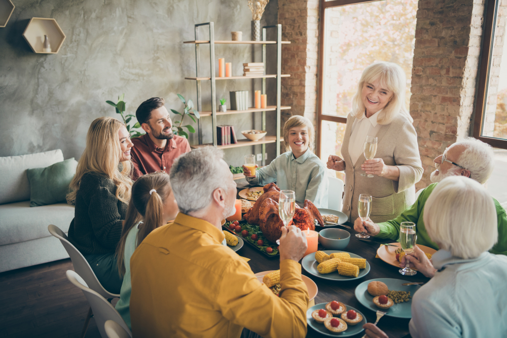 A family of eight, including children and older adults, gathers around a table with a roast turkey and other dishes, smiling and raising glasses in a bright, cozy dining room.