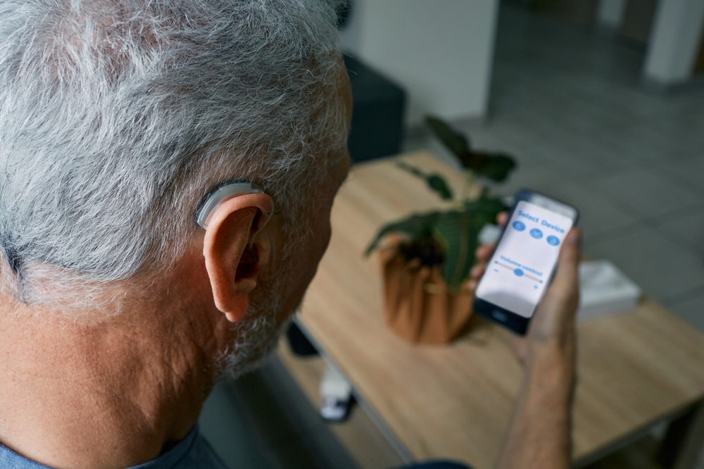 An older man with gray hair adjusts the settings on his smartphone for his behind-the-ear hearing aid while sitting at a table with a potted plant.