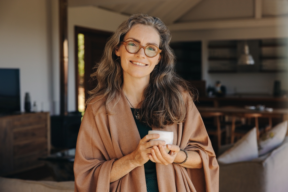 A woman with long gray hair and glasses, wearing a brown shawl, stands indoors holding a white mug and smiling at the camera. The background shows a cozy, modern living space with wooden furniture.
