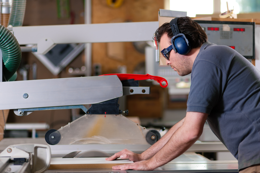 A man wearing safety glasses and earmuffs operates a circular saw in a workshop, guiding a piece of wood through the machine with both hands.