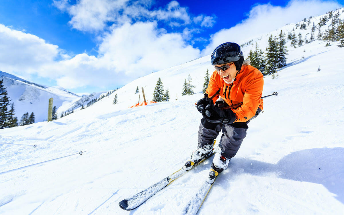 A person in an orange jacket and black helmet skis down a snowy mountain slope, smiling, with pine trees and a bright blue sky in the background.