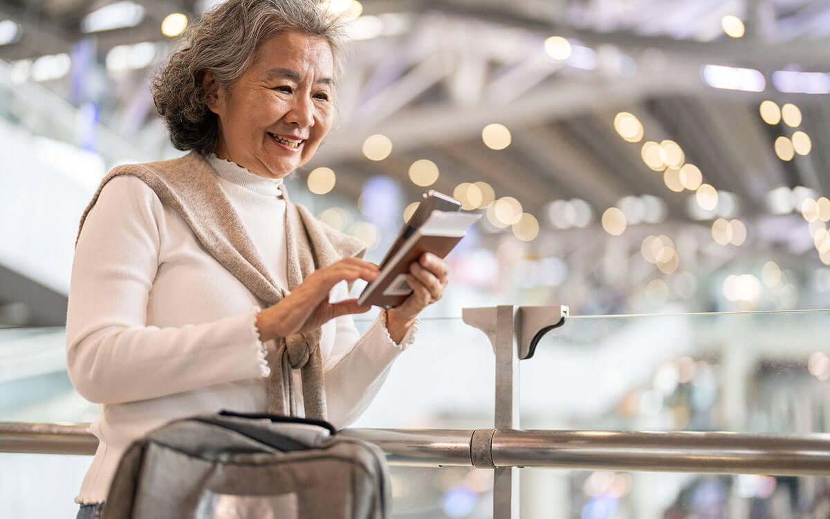 An older woman smiling while holding a smartphone and a passport at an airport, with a soft focus on the bright, modern terminal in the background. She has a sweater over her shoulders and a bag in front of her.