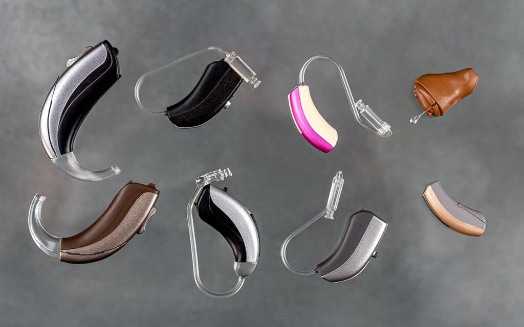 Eight different hearing aids in various colors and shapes are arranged on a gray background, showcasing a variety of styles and designs.
