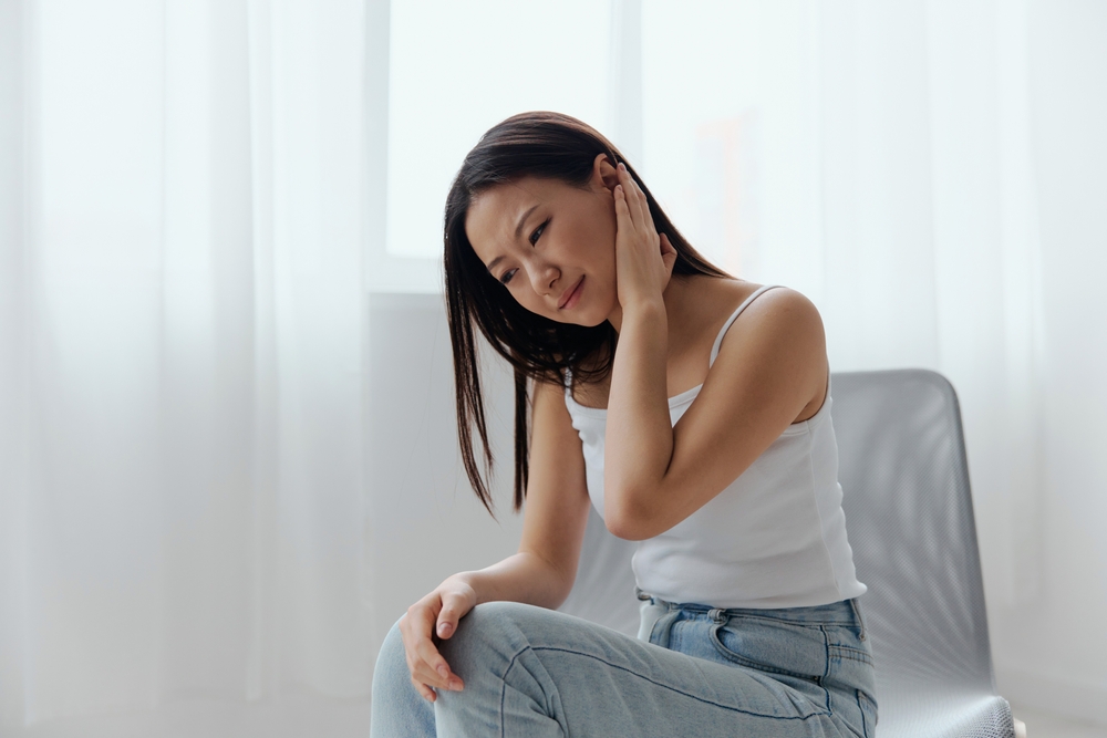 A woman in a white tank top and jeans sits on a chair, tilting her head and touching her ear with a pained expression, suggesting she may have ear discomfort or pain. Light curtains are in the background.