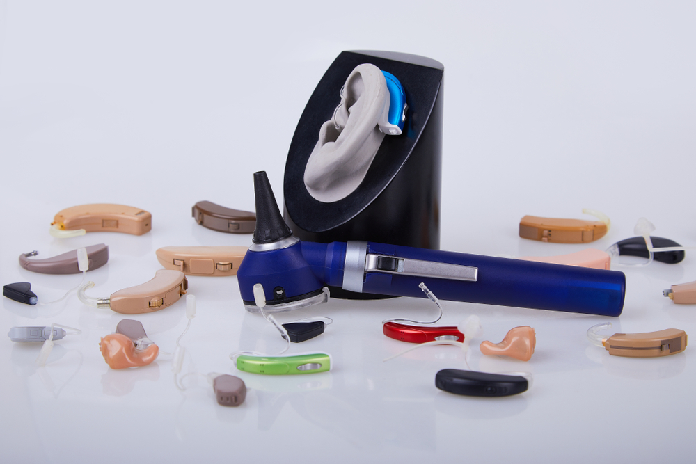 Various hearing aids in different colors and styles are scattered on a white surface, with an otoscope and a model ear displaying a blue hearing aid placed prominently in the center.
