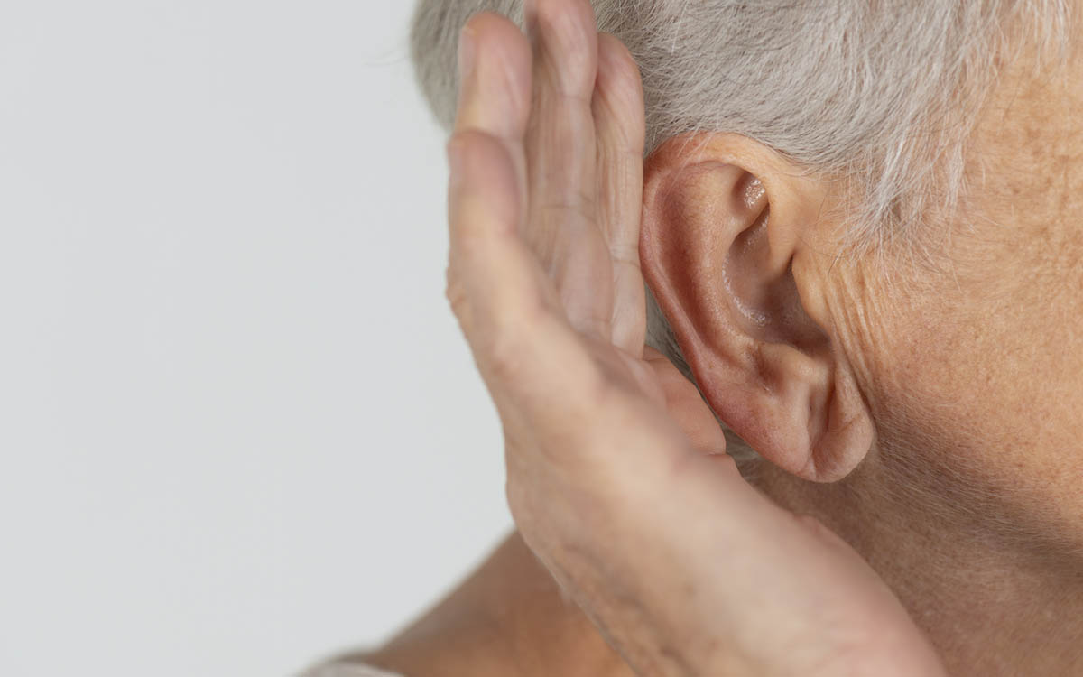 Close-up of an older adult holding a hand to their ear, suggesting difficulty hearing or trying to listen more closely. The background is plain and light-colored.