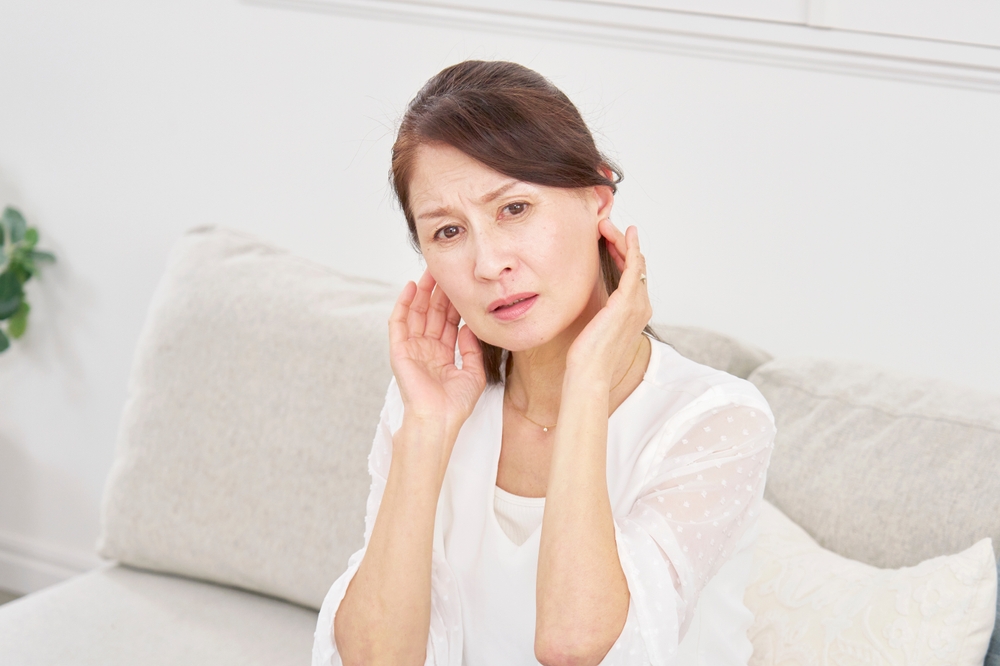 A middle-aged woman sits on a sofa, touching both of her ears with a concerned expression, suggesting she may be experiencing ear discomfort or pain.