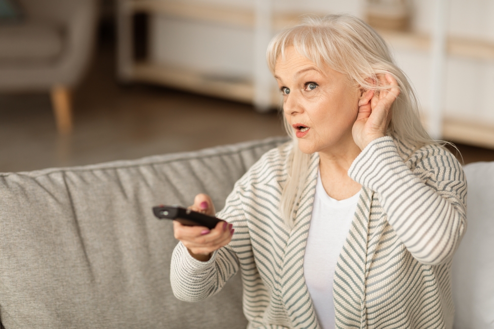 An older woman sits on a couch holding a TV remote, raising her other hand to her ear as if trying to hear better, with a concerned expression on her face.