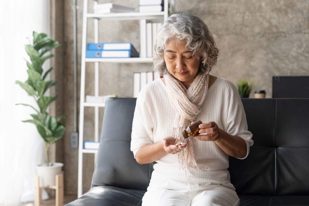 An older woman with gray hair sits on a black couch, looking down as she pours pills from a brown bottle into her hand. She wears a light scarf and white clothes. A bookshelf and plant are in the background.