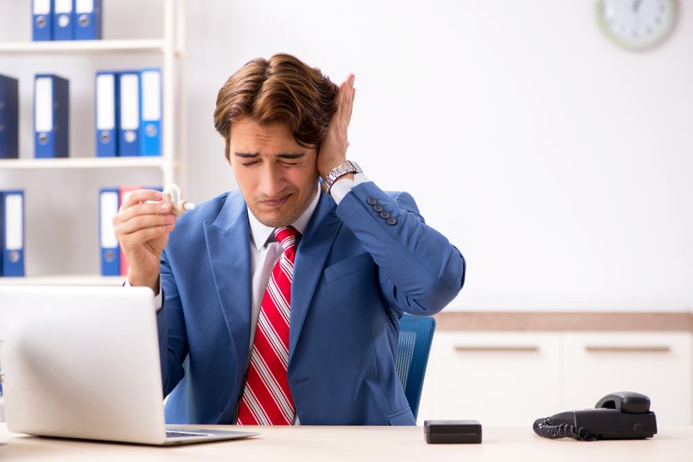 A man in a suit sits at a desk with a laptop, holding a cup and covering one ear, looking uncomfortable as if disturbed by a loud noise. A phone, notebook, and office shelves are visible in the background.