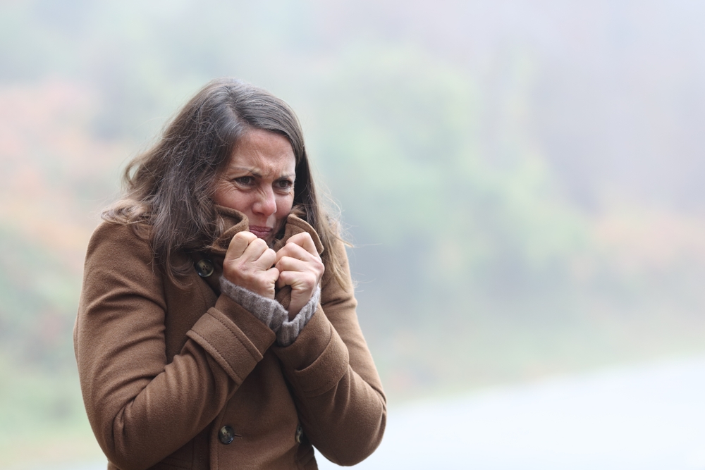 A woman in a brown coat stands outside, shivering and holding her collar close for warmth. The background is blurred with green foliage and mist, indicating cold weather.