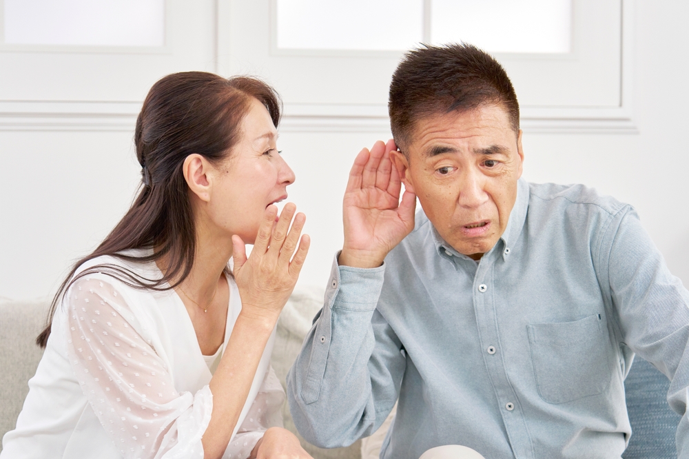 A woman whispers into the ear of a man who is cupping his ear and leaning in, appearing to have difficulty hearing her. They are sitting together indoors in a bright room.