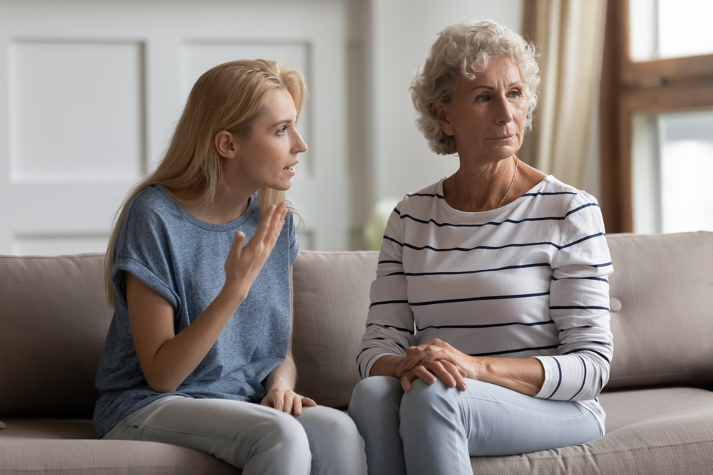 A younger woman is talking to an older woman on a couch. The younger woman appears upset or frustrated, while the older woman looks away with a serious or distant expression. Both are indoors in a bright room.