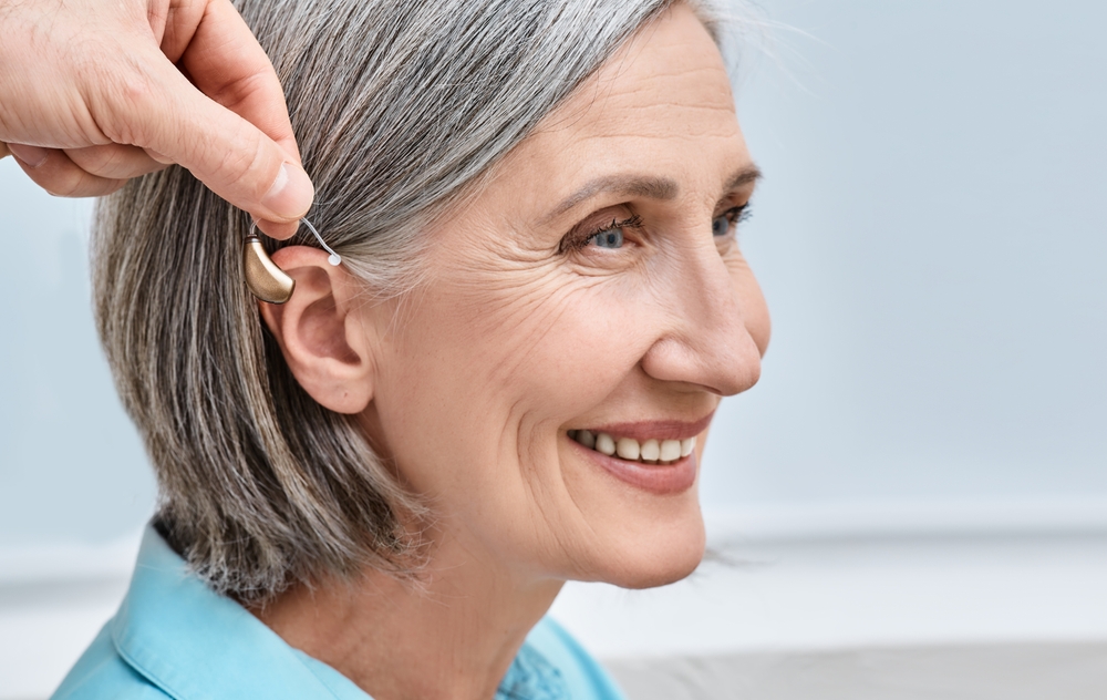 A smiling older woman with gray hair is having a hearing aid placed behind her ear by another persons hand.
