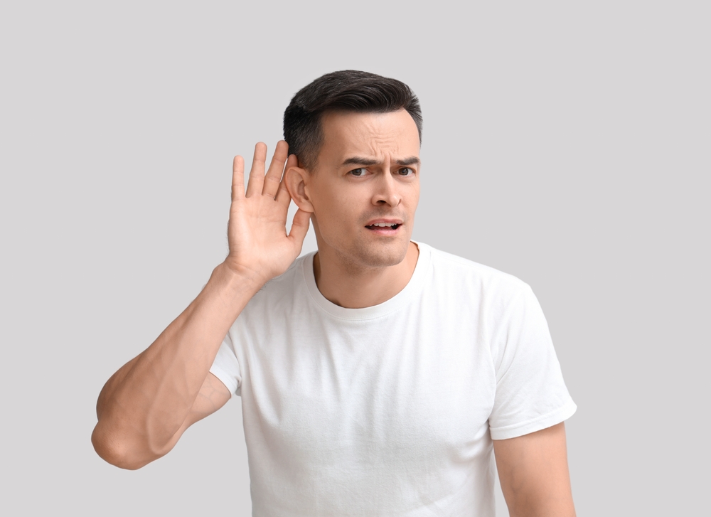 Man in a white t-shirt holding his hand to his ear, tilting his head, and looking slightly confused as if trying to hear something better, against a plain light gray background.