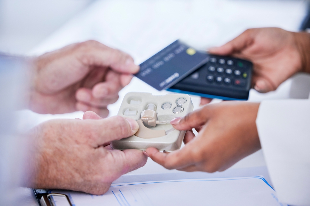 A person pays with a credit card using a card reader held by someone else, while receiving a hearing aid device in a tray.