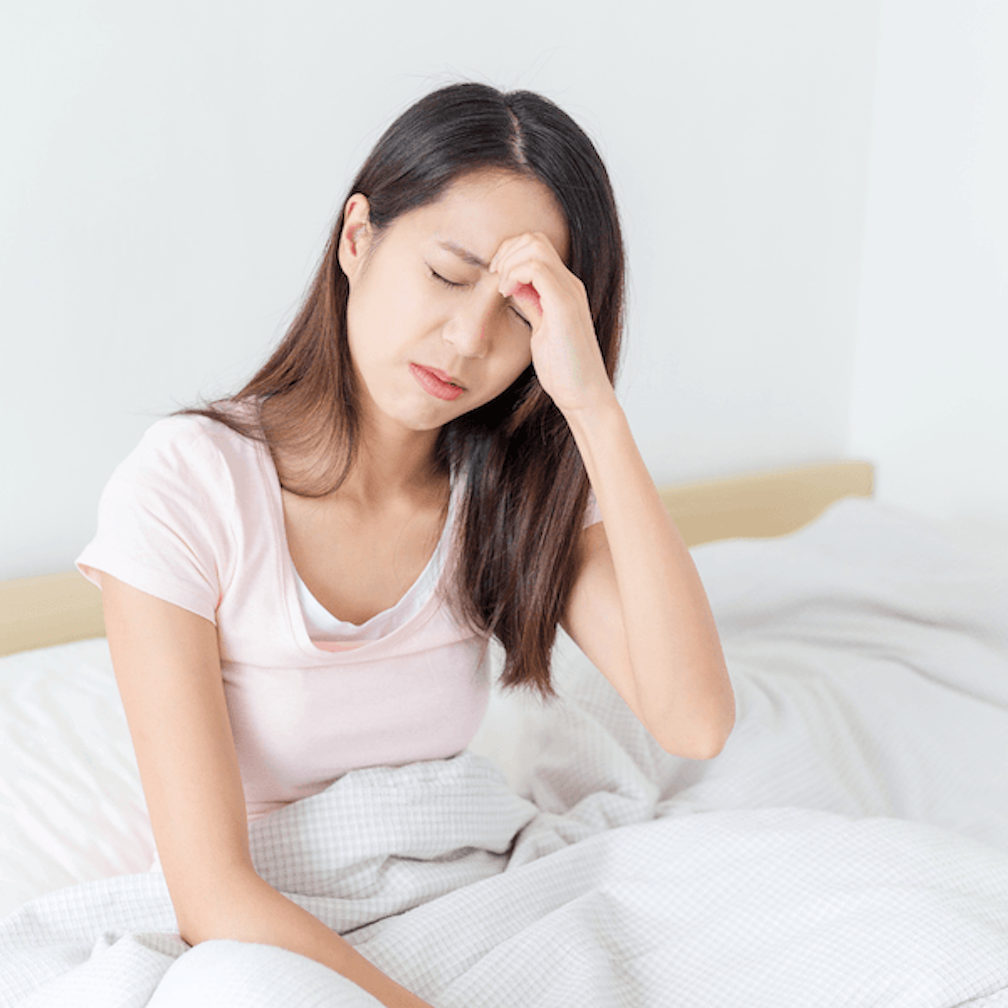 A woman sits on a bed with her eyes closed, touching her forehead as if she has a headache or is feeling unwell. She is covered with a white blanket and looks visibly uncomfortable.