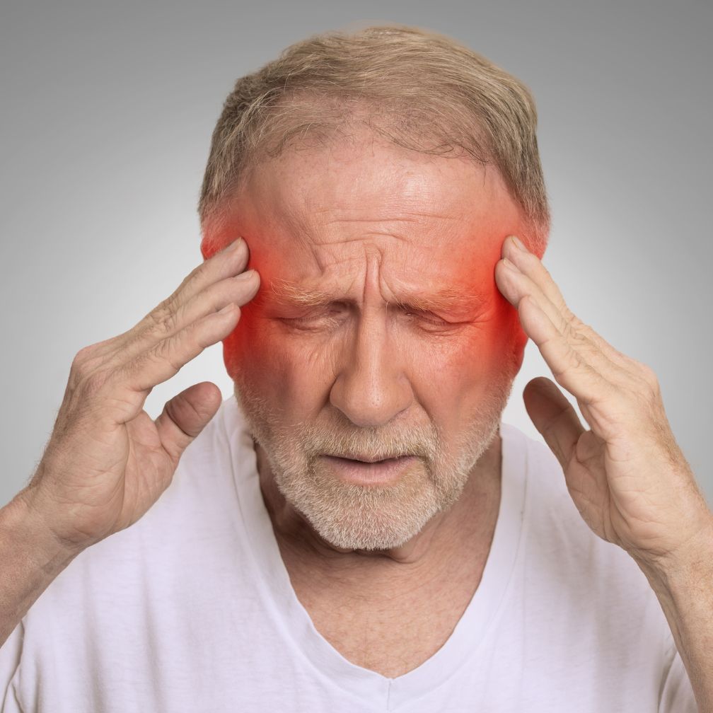 Older man holding his temples with both hands, eyes closed, and a pained expression. Red coloring on his temples indicates head pain or a headache. He is wearing a white shirt against a plain gray background.