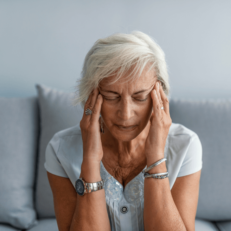 An older woman with short gray hair sits on a couch with her eyes closed, touching her temples as if she has a headache or is stressed. She wears a watch, rings, bracelets, and a light blue blouse.