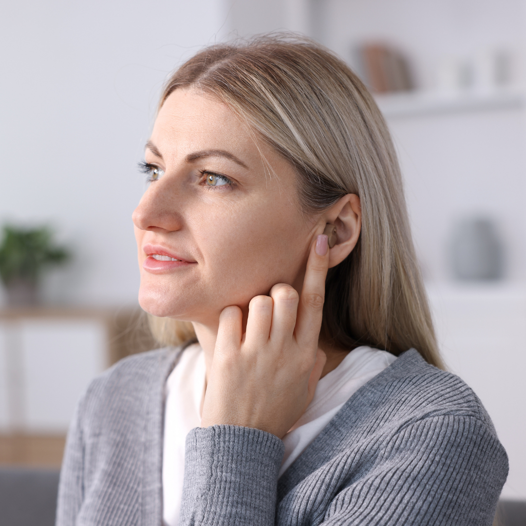 A woman with blonde hair touches her ear with her finger, appearing thoughtful. She is indoors, wearing a gray sweater, with a blurred background featuring shelves and a plant.