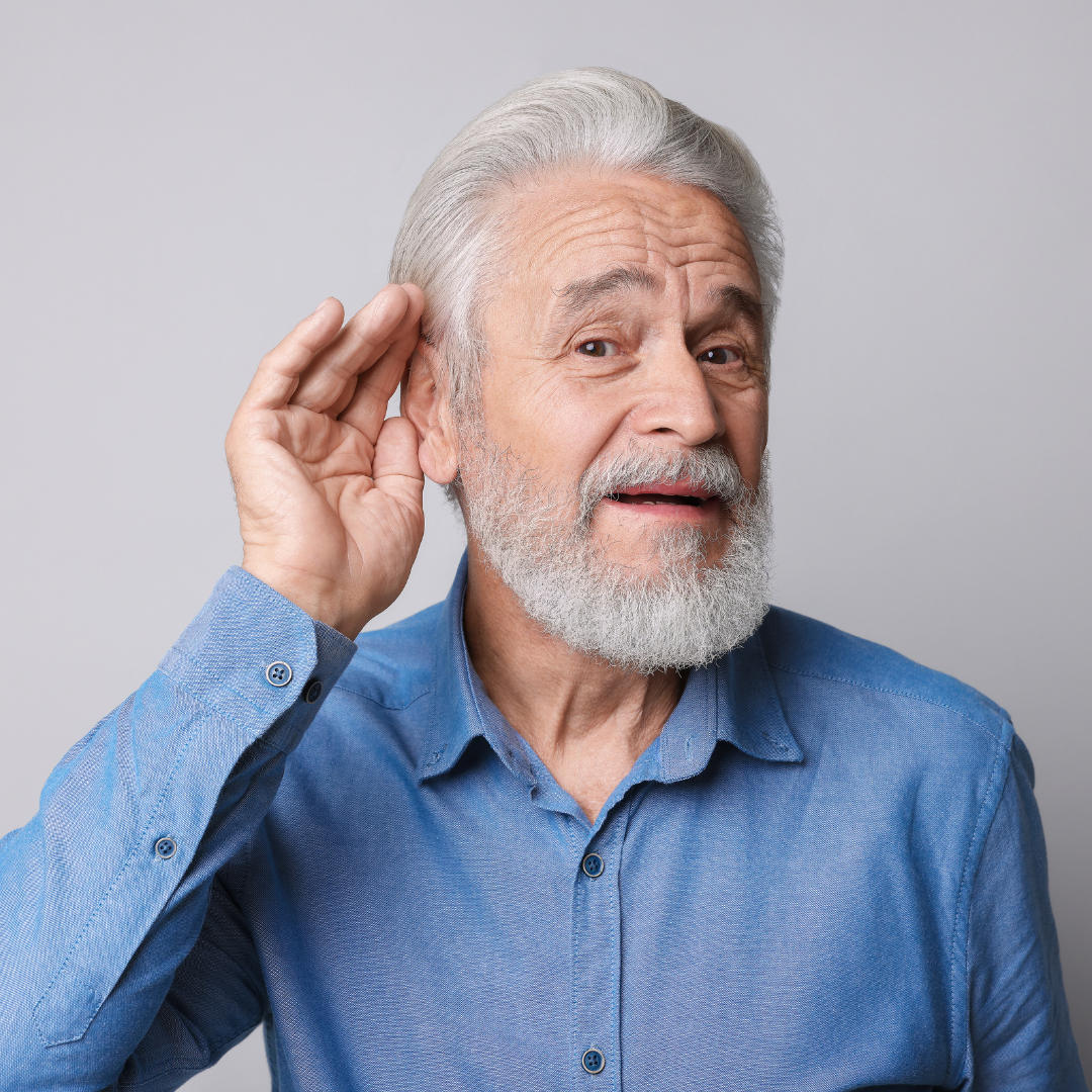 Older man with gray hair and beard, wearing a blue shirt, tilting his head and holding a hand to his ear as if trying to hear better, against a plain light background.